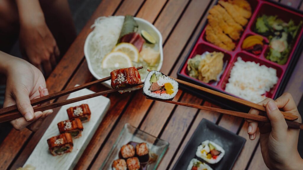 Close-up of hands holding sushi with chopsticks, showcasing Japanese cuisine (bento box) over a wooden table.