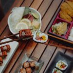 Close-up of hands holding sushi with chopsticks, showcasing Japanese cuisine (bento box) over a wooden table.