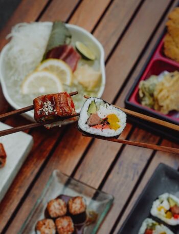 Close-up of hands holding sushi with chopsticks, showcasing Japanese cuisine (bento box) over a wooden table.