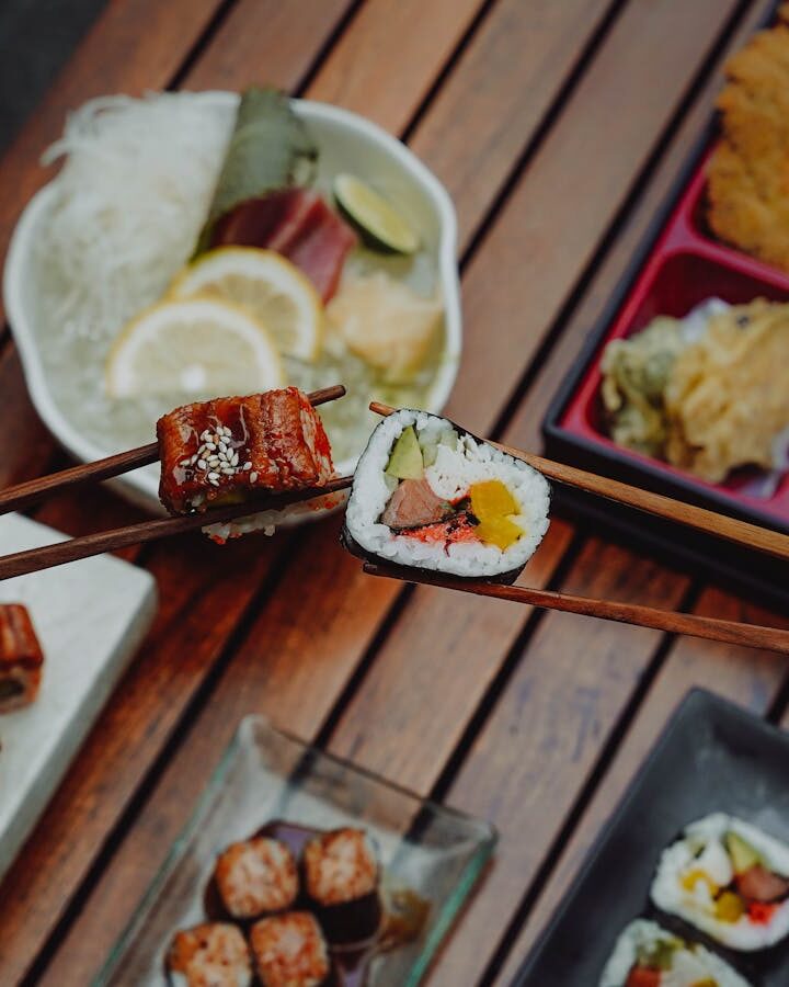Close-up of hands holding sushi with chopsticks, showcasing Japanese cuisine (bento box) over a wooden table.