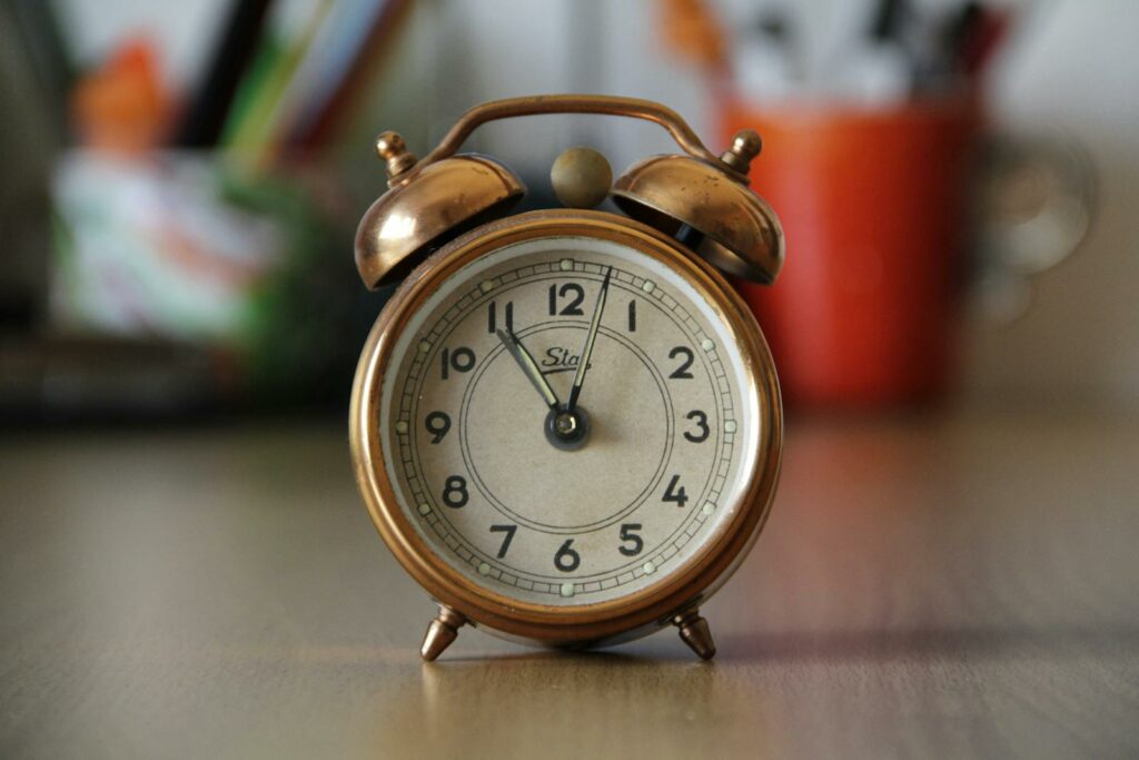 A vintage copper alarm clock with bells placed on a desk with blurred objects in the background.